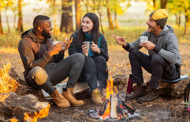 Three people sit round a campfire drinking from mugs