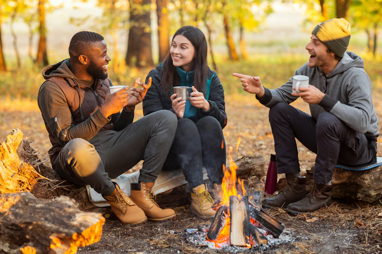 Three people sit round a campfire drinking from mugs