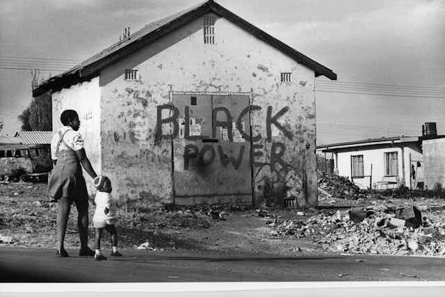 A woman holding a child's hand as they walk past a building with the graffiti: Black Power.