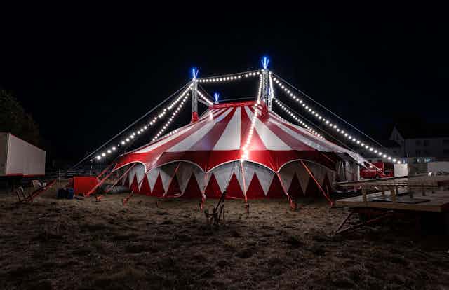 A red and white circus tent in a field at night, lit up with small white strings of lights.