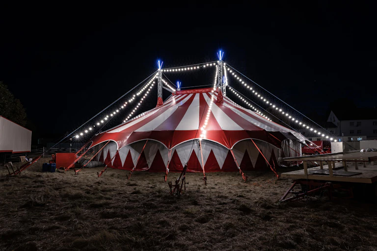 A red and white circus tent in a field at night, lit up with small white strings of lights.
