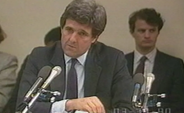 Screen grab from grainy video of man in suit seated before a microphone flanked by a young woman and a young man.