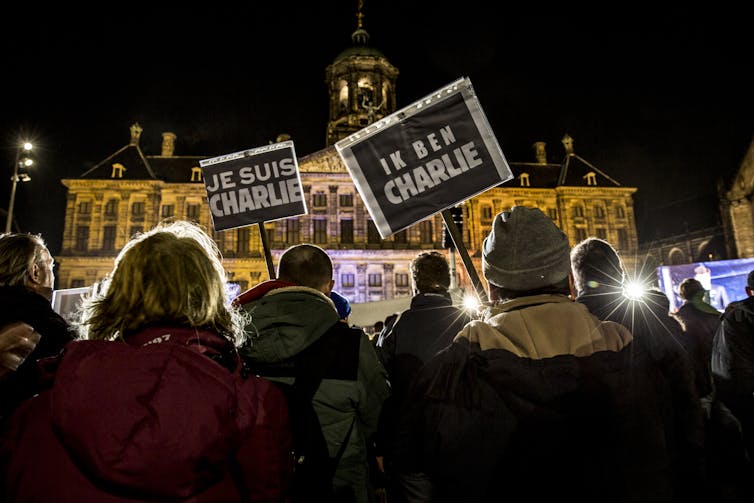 A crowd of demonstrators at night in front of a public building in Amsterdam, holding signs that say Je Suis Charlie and Ik Ben Charlie