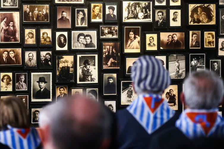 Four people seen from the back stand facing a black wall covered in old black-and-white photo portraits.