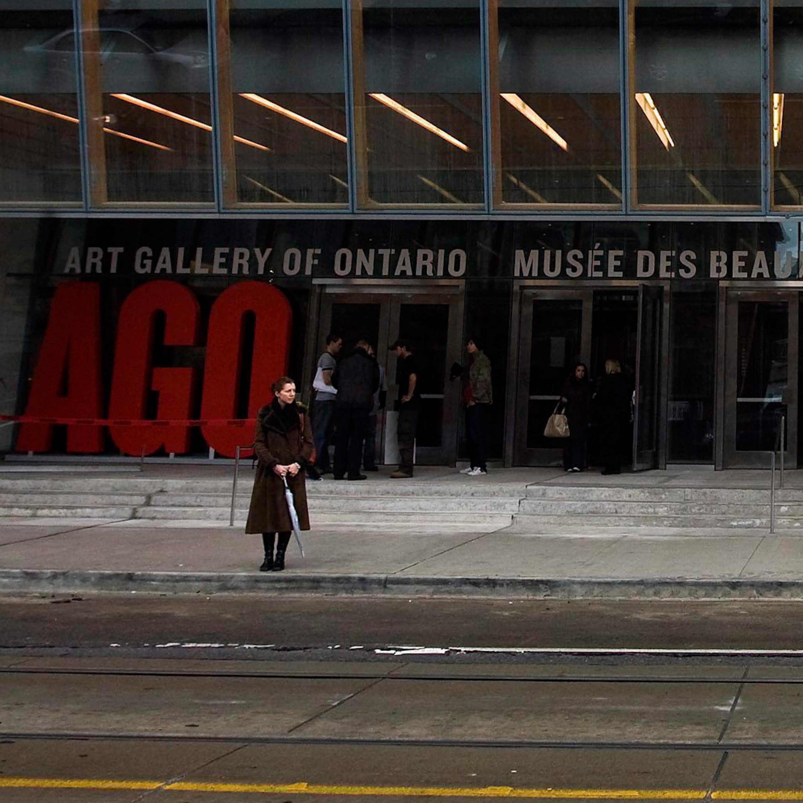 A person with umbrella seen in front of a building with wide steps and many glass panes that says Art Gallery of Ontario.