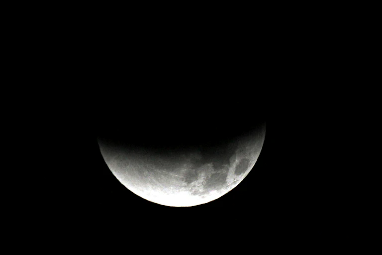 The moon, partially shadowed on the top side, against a dark background.