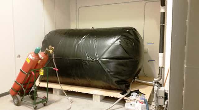 A large, cylindrical bag inflated and laying on its side on a wooden pallet, connected to two cylindrilcal tanks on a wheeled cart. 