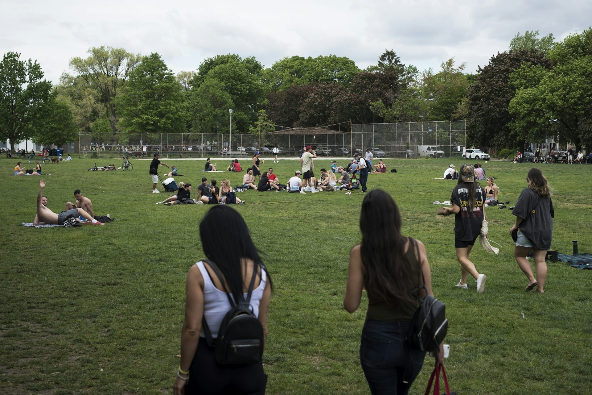 People in a large green space with a baseball diamond in the background and two young women seen from behind in the foreground