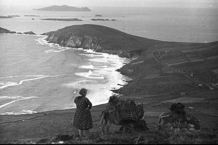 Black and white photo of a woman standing in front of a sweeping sea view with a donkey.