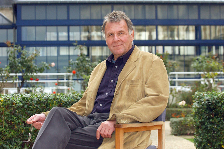 A middle-aged man in a beige corduroy jacket sitting on a chair in a rose garden.
