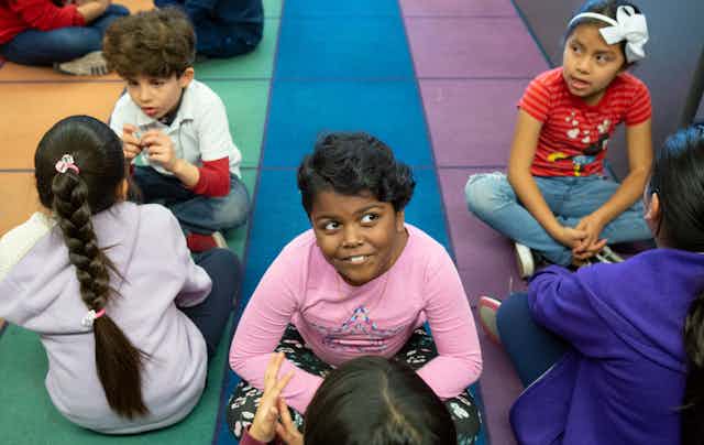 Students seen sitting on tiles in a classroom and one girl is smiling and pensive.