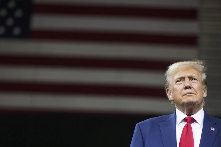 A man in a coat and tie stands in front of an American flag.