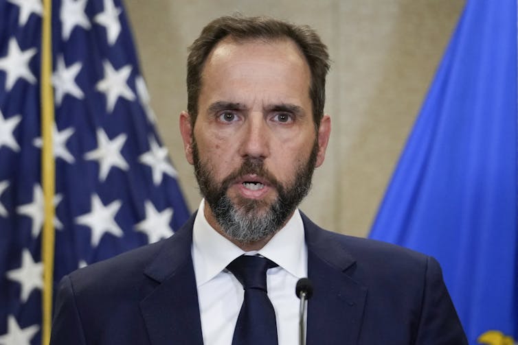 A bearded man in a coat and tie stands in front of an American flag.