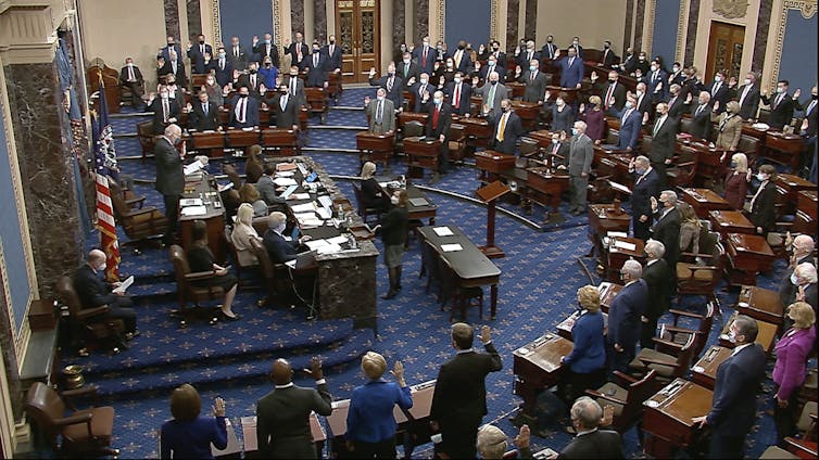 Una vista de una cámara legislativa formal con muchas personas paradas en sus escritorios y en un estrado.