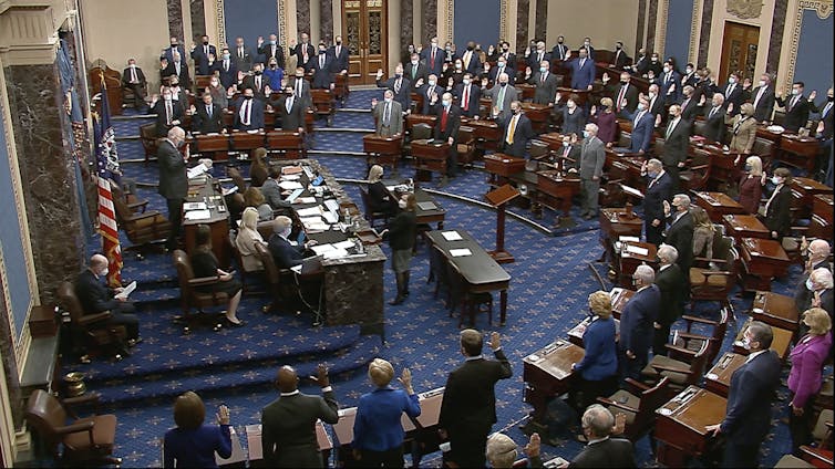 A view of a formal legislative chamber with many people standing at their desks and on a dais.