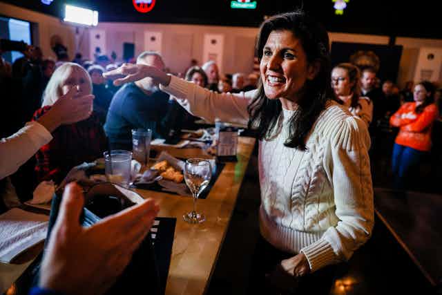 Nikki Haley wears a beige sweater and stands behind a bar, with people seated at it. She smiles and appears ready to shake one person's hand. 