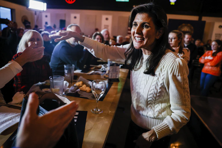 Nikki Haley wears a beige sweater and stands behind a bar, with people seated at it. She smiles and appears ready to shake one person's hand.