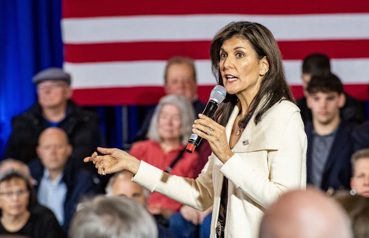 Nikki Haley wears a white jacket and stands in front of a group of seated people, with the backdrop of the American flag. She holds a microphone and points her finger towards the crowd.
