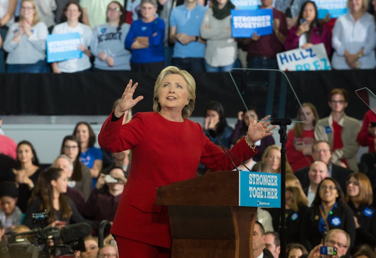 Hillary Clinton wears a red pantsuit and gestures while standing at a podium, in front of a large crowd of people.