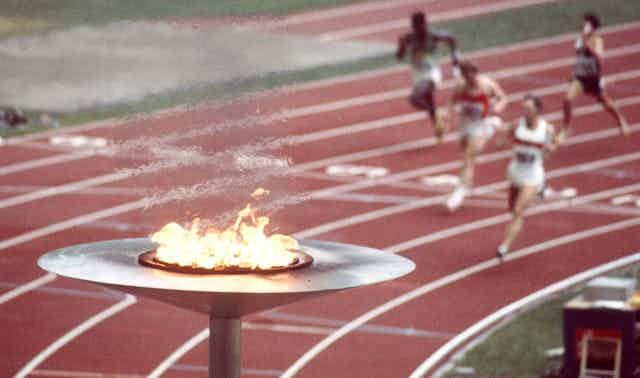 A large torch burning in the foreground while track athletes run in the background