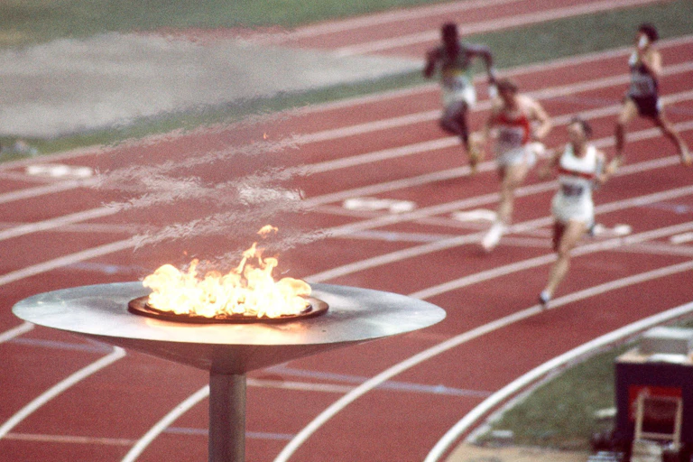 A large torch burning in the foreground while track athletes run in the background