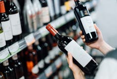 In an over-the-shoulder photograph, a woman chooses between two bottles of wine at a liquor store.