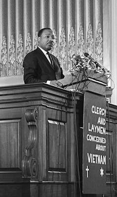 A Black man wearing a dark suit stands behind a lecturn atop a sign that says clergy and laymen concerned about Vietnam.