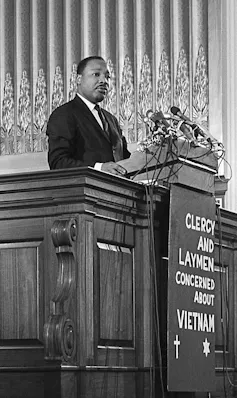A Black man wearing a dark suit stands behind a lecturn atop a sign that says clergy and laymen concerned about Vietnam.