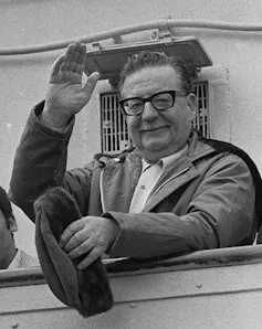 A black and white photo of a man wearing glasses waving