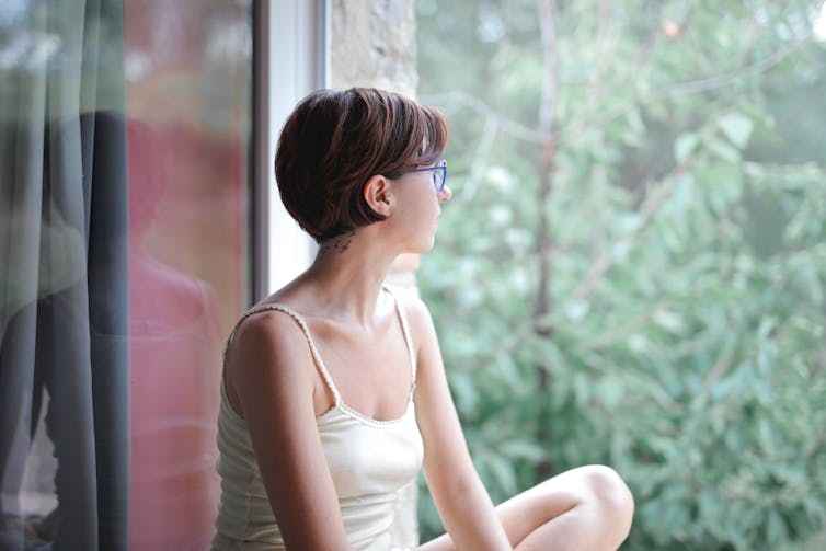 A woman sits by a window gazing out onto trees outside.