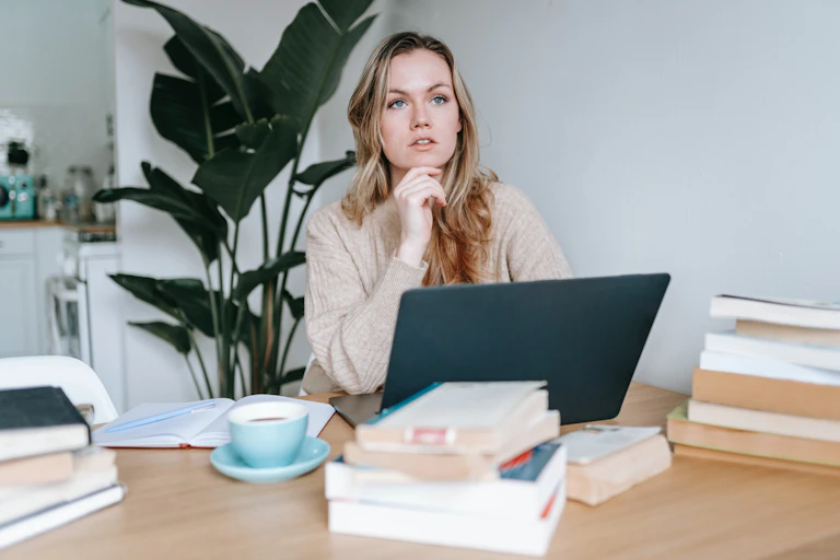 A woman working at a table crowded with books looks away from her laptop screen.
