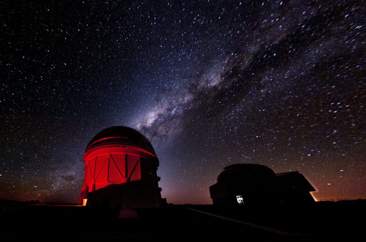 Foto de um prédio de observatório iluminado em vermelho com o céu estrelado ao fundo.