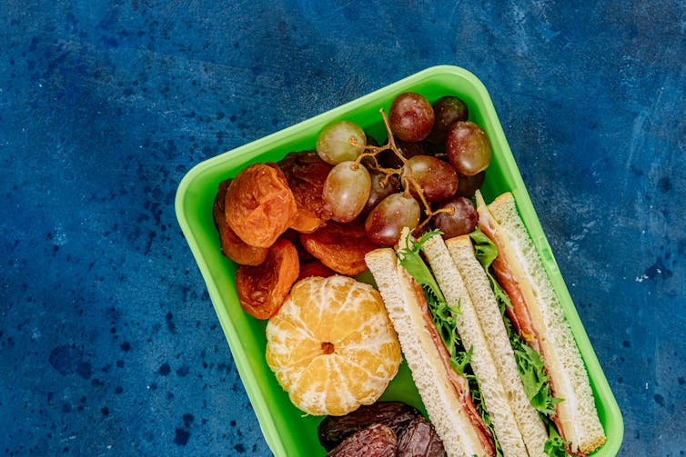 A lunch box with a peeled mandarin, grapes, dried apricots and a sandwich.
