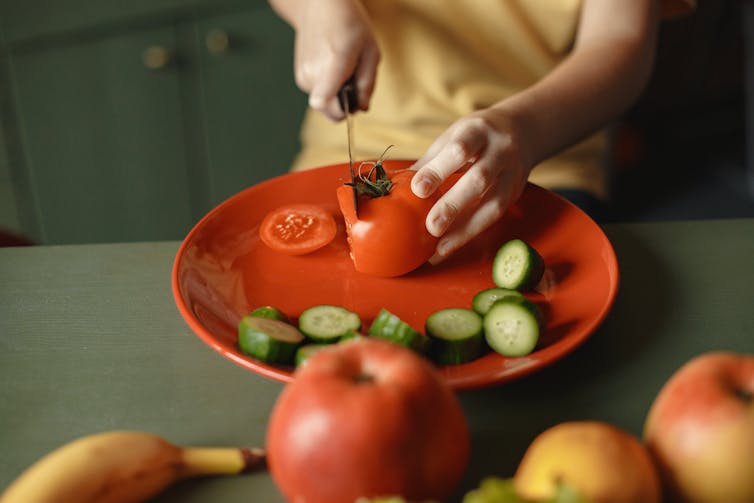 A young child chops tomato on a plate with chopped cucumbers.