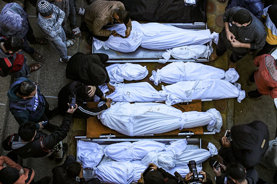 Aerial view of human forms wrapped in white cloth on stretchers, with people standing around them.