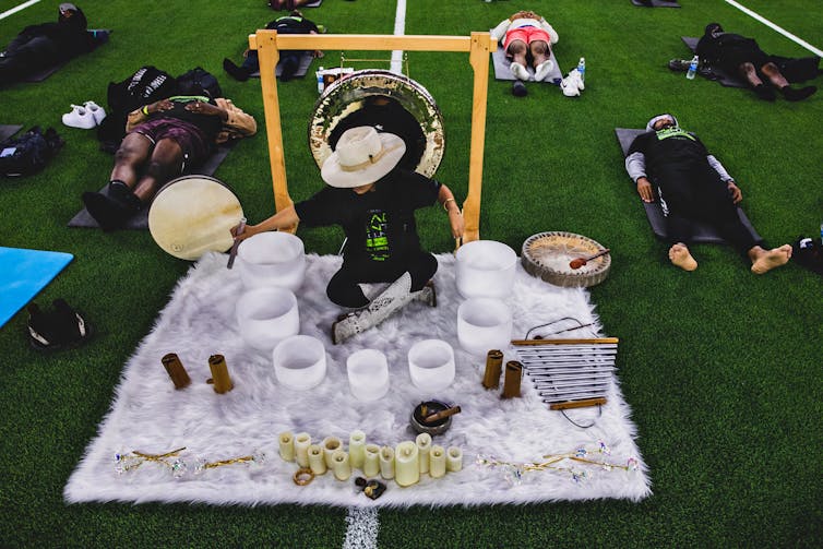 A person in a black outfit and white hat playing sounds on singing bowls while several others lie in meditation poses nearby.