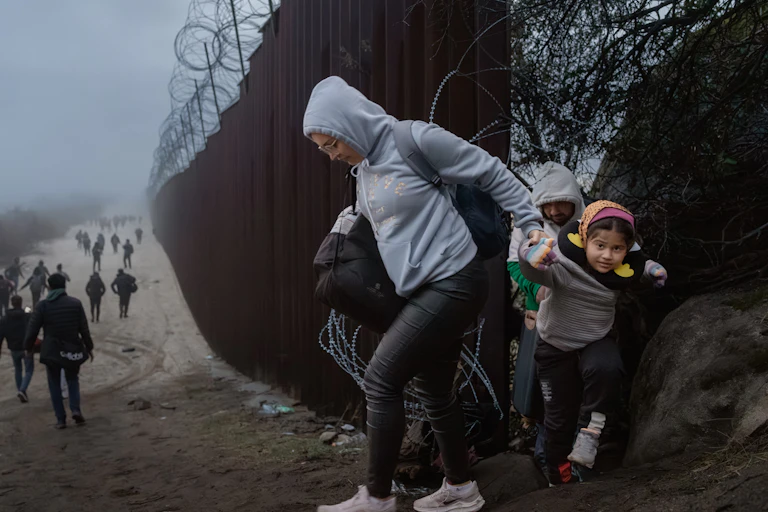 A woman wearing a grey sweatshirt pulls a child through a role in a dark fence, with barbed wire on the top. Other people walk along the fence in the distance.
