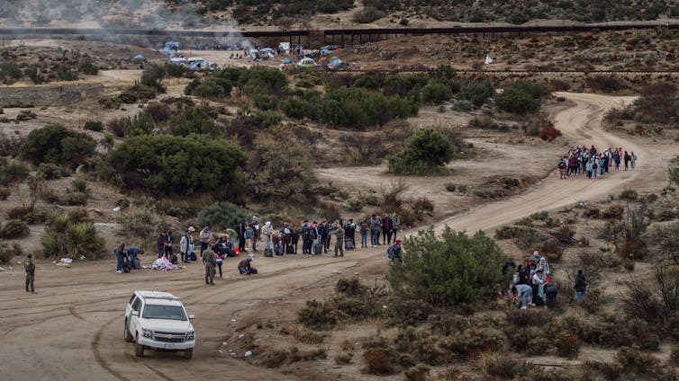 People are seen standing in a desert on a grey day with a white SUV nearby.