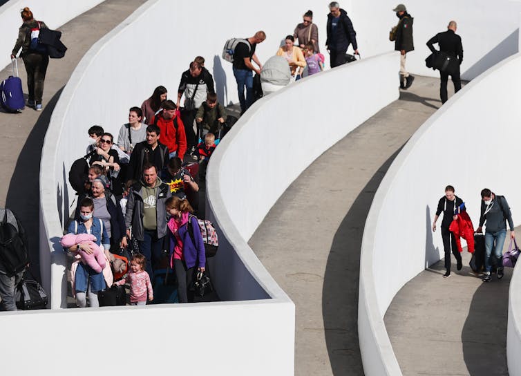 People walk along white walls that wrap in a circular manner. The people carry backpacks, children and wear jackets.
