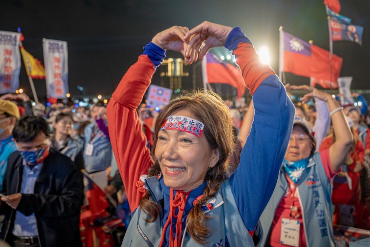 A woman makes a heart shape with her arms, behind her are people carrying flags and placards.