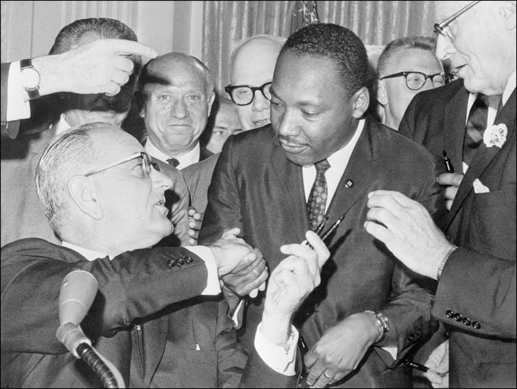 A white man is shaking the hands of a Black man as a crowd of other men stand behind them.