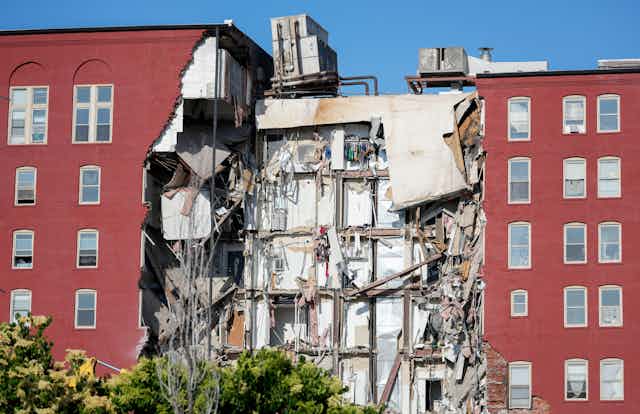 Several floors of a red brick six-story apartment building are gone. Clothing is still visible in closets.