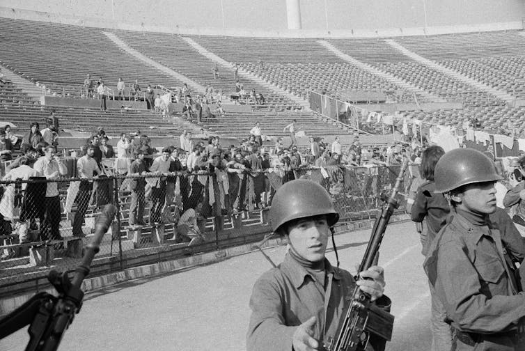 a black and white photo of soldier guarding prisoners in a stadium