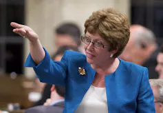 A woman with short dark hair and glasses gestures as she speaks.