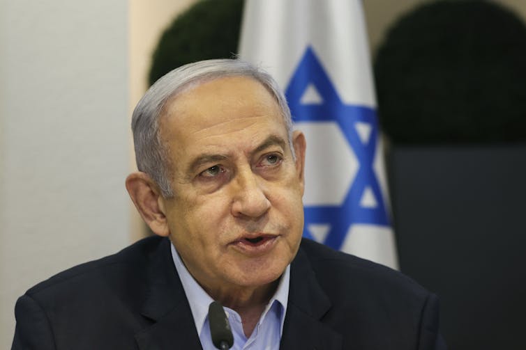 A man with grey hair talks in front of an Israeli flag.