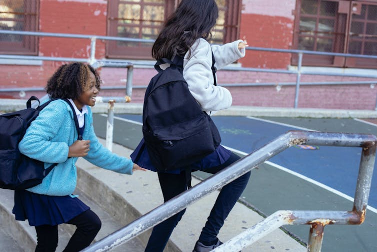 Two high school students carrying backpacks on one shoulder walk up steps in a playground.