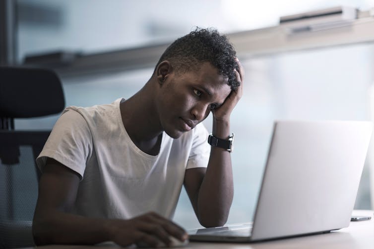 A young black man looks at a laptop screen with a sad expression