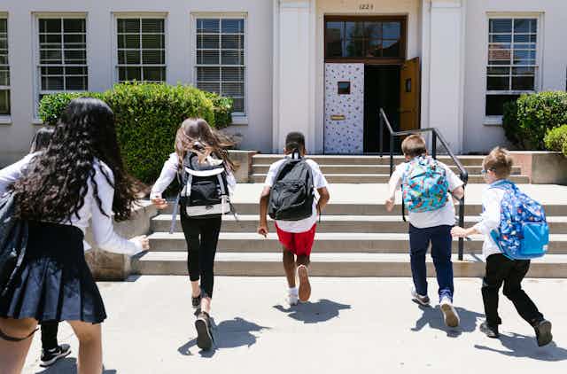 Six school students, wearing back packs run into a school building. 