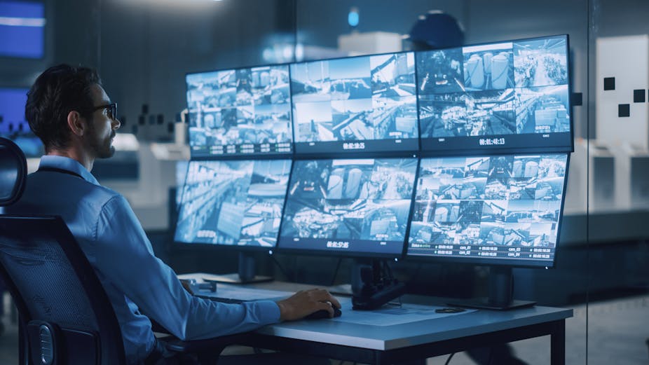  A man wearing spectacles looking at images on six computer monitors.