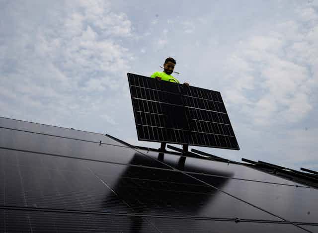 A man holds up a solar panel on a roof against a cloudy sky.